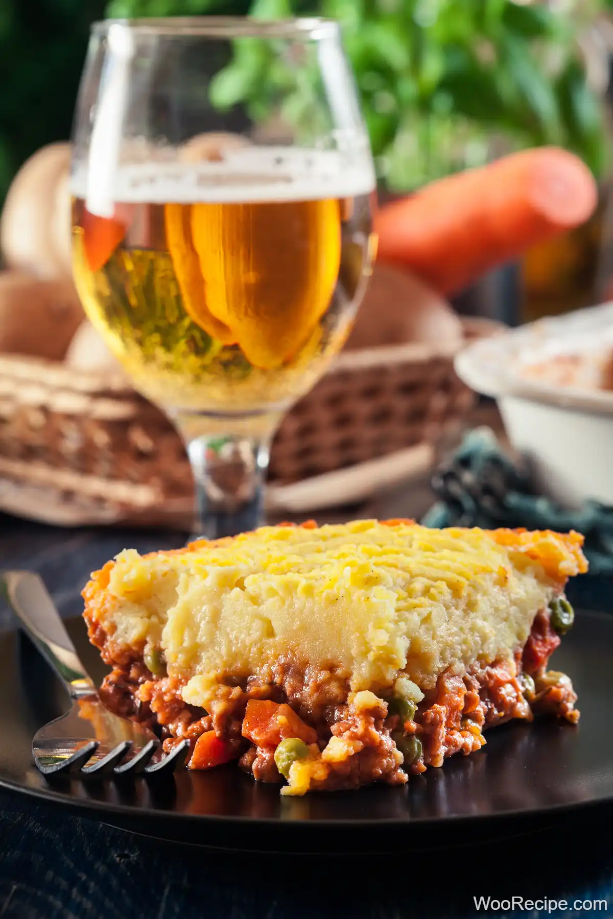 A slice of traditional English shepherd's pie rests on a black plate with a fork beside it. The dish boasts a creamy layer of mashed potatoes over a savory meat and vegetable filling. A glass of beer and fresh vegetables blur softly in the background, adding to the cozy ambiance.