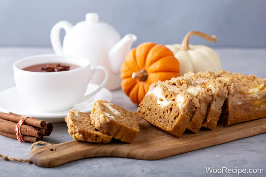 A wooden board showcases pumpkin bread with a luscious cream cheese filling, accompanied by cinnamon sticks. In the background, a cup of tea mingles with a white teapot and small orange and white pumpkins.