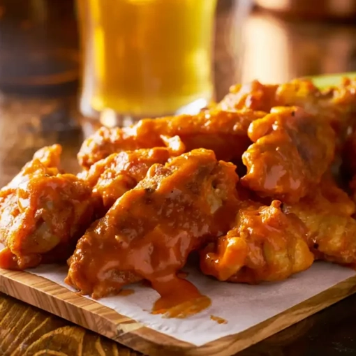 A wooden tray holds a pile of saucy buffalo chicken wings glistening with a rich orange-red glaze In the blurred background theres a pint of beer creating a cozy and inviting atmosphere