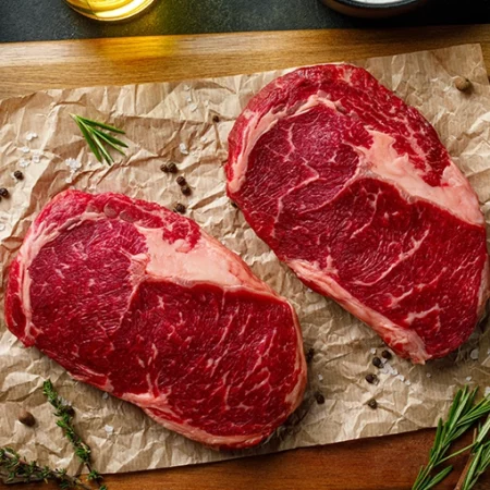 Two raw beef ribeye steaks rest on crumpled parchment paper surrounded by sprigs of rosemary and scattered peppercorns The marbled steaks sit upon a wooden board with a small bottle of oil partially visible in the backdrop