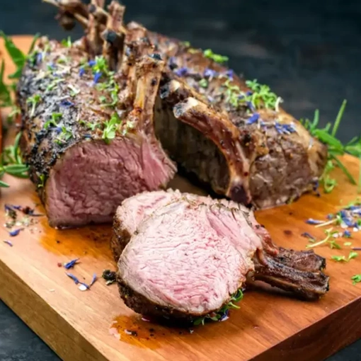 A roasted rack of lamb sits on a wooden cutting board garnished with fresh herbs and small flowers One chop is sliced to show the pink juicy interior revealing a perfectly cooked piece of meat
