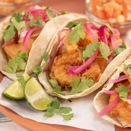 Three Mexican tacos filled with fried fish pickled red onions and fresh cilantro are displayed on a tray Lime wedges and a side of salsa are visible in the background