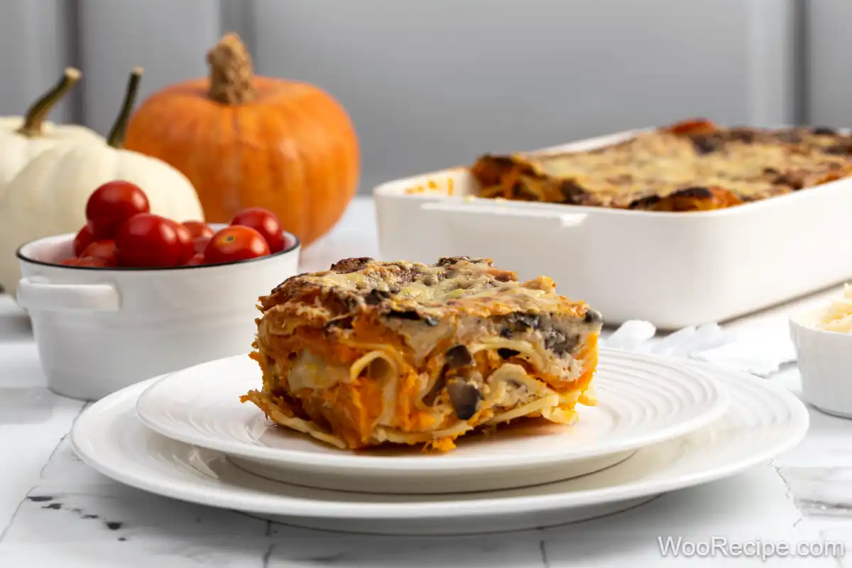 A delicious slice of homemade pumpkin lasagna served on a white plate, surrounded by fresh cherry tomatoes and a seasonal pumpkin. The creamy layers of cheesy pumpkin goodness are beautifully visible, showcasing a perfect autumn dish ideal for comforting meals. A baking dish of lasagna sits in the background, ready to be served, creating a warm and inviting atmosphere perfect for family gatherings.