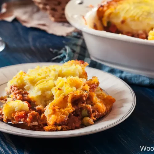 A comforting plate of Traditional English Shepherd's Pie, featuring a golden-brown layer of mashed potatoes atop a savory mixture of ground meat and vegetables. The dish is served on a decorative plate with a fork resting nearby, evoking cozy family meals and hearty flavors. In the background, a baking dish of Shepherd's Pie awaits serving.