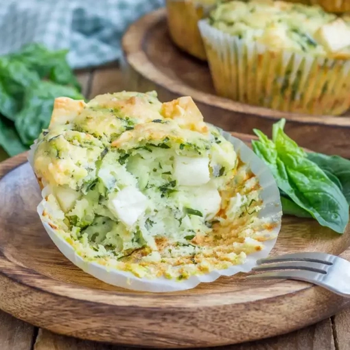 A savory muffin with visible spinach and cheese pieces partially unwrapped and placed on a wooden plate Fresh spinach leaves are next to the muffin with a fork nearby resting on a rustic wooden table